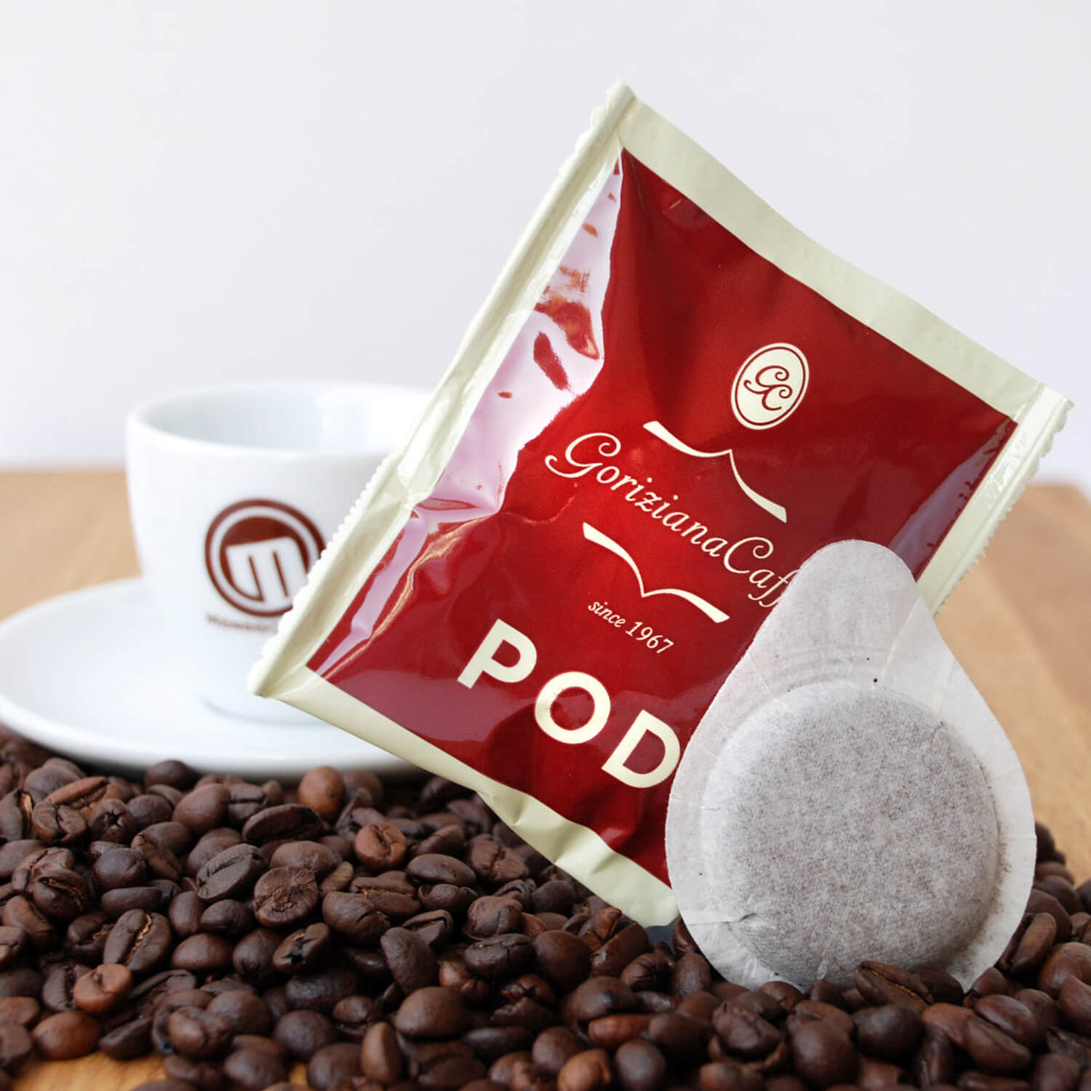 Eine Packung Torrefazione Goriziana Espresso ESE Pads (150 x 7 g) mit einer Tasse und Untertasse, umgeben von Kaffeebohnen auf einer Holzfläche.