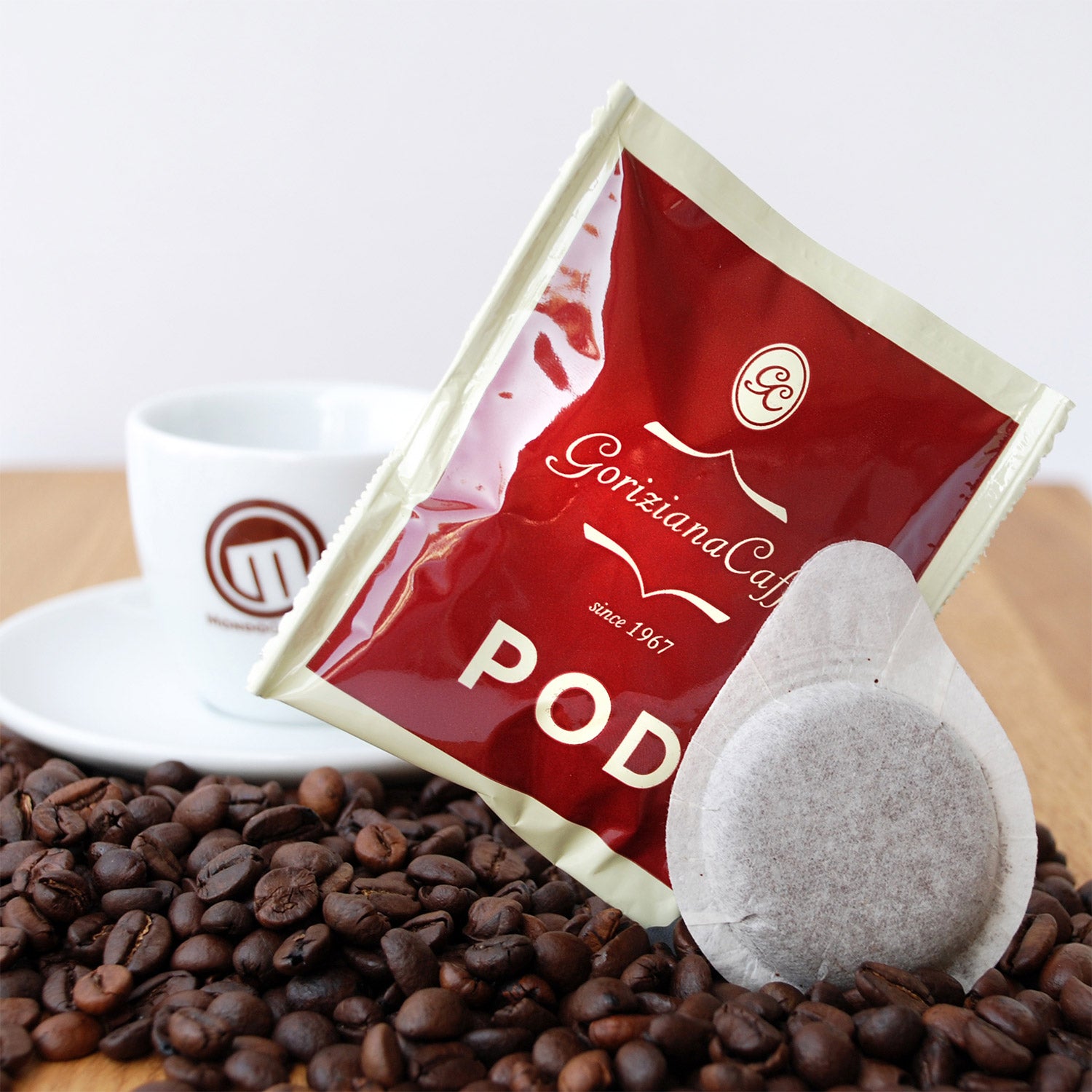 Torrefazione Goriziana Espresso ESE Pads (15 x 7g) und eine Kapsel mit Kaffeebohnen, neben einer weißen Tasse und Untertasse auf einer Holzunterlage platziert.