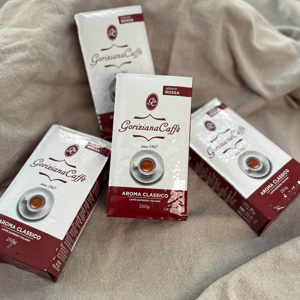 Vier Packungen Torrefazione Goriziana Espresso gemahlen ROSSA Aroma Classico, 4 x 250 g, auf beigefarbenem Stoffgrund.