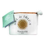 Ein weißer Keramikbehälter mit der Aufschrift Sal de Ibiza GmbH, der 140 g Fleur de Sel - Isla blanca mit ibizenkischen Kräutern enthält, wird mit einem Korkdeckel, einem Keramiklöffel und einem grünen Anhänger geliefert.