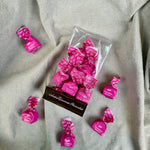 Eine durchsichtige Schachtel mit Viani & Co. Tartufini dolci bianchi - weiße Mini Schokoladentrüffel, 100 g, und mehrere rosa verpackte Pralinen werden auf einem hellgrauen, strukturierten Stoffhintergrund präsentiert.