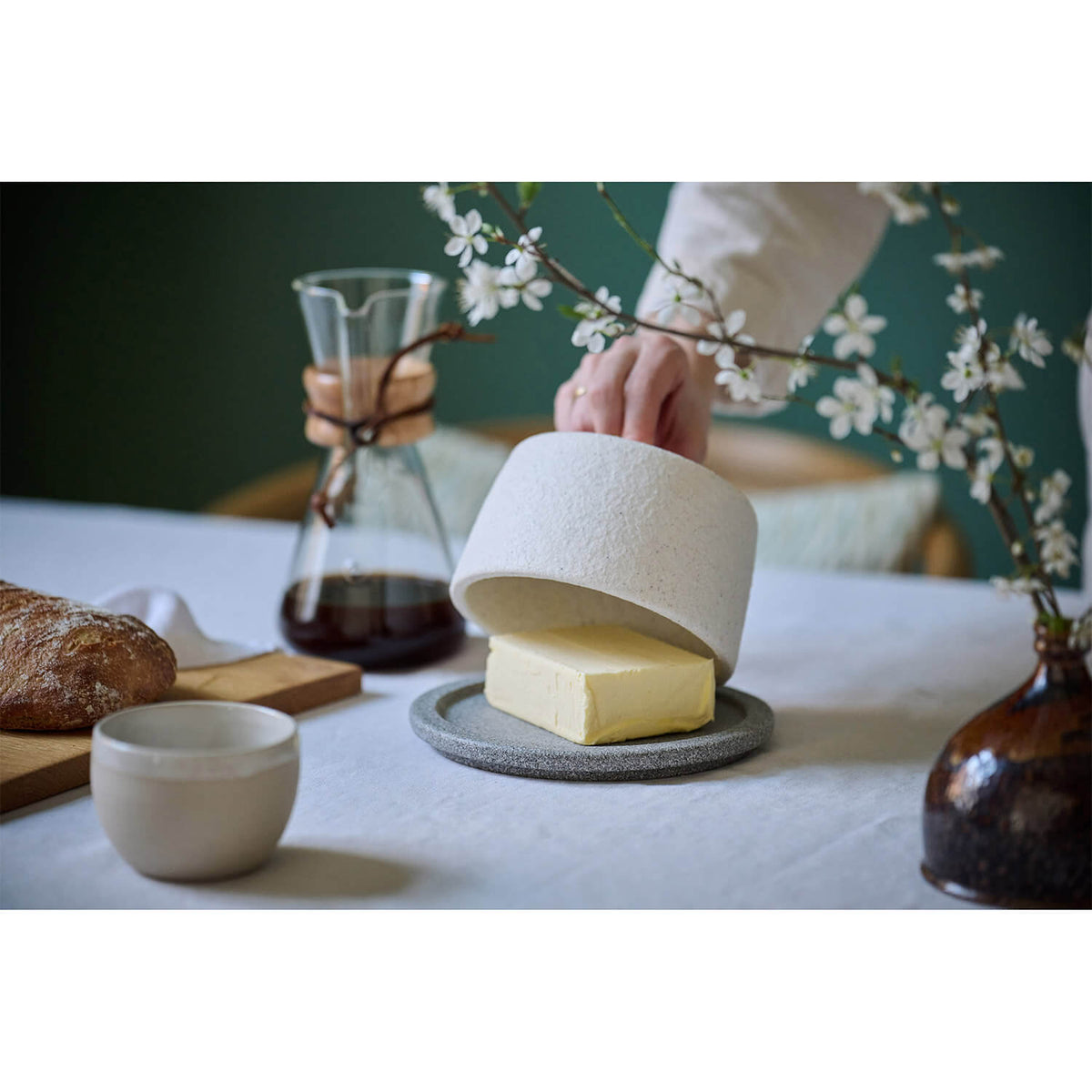 Eine Hand hebt den Butterkühler von Denk by Denk Keramik an und enthüllt Butter auf einem Teller; Kaffee, Brot und Blumen stehen auf dem Tisch.